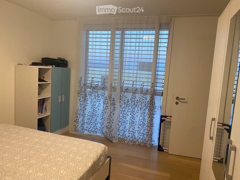 A tidy bedroom with a bed, a white cabinet with shelves, a printer on top, and sheer curtains with a leafy pattern covering the windows.