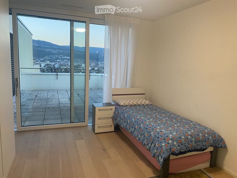 Cozy bedroom with a single bed, nightstand, and drawer unit. Large sliding glass door opens to a balcony with a scenic city and mountain view.