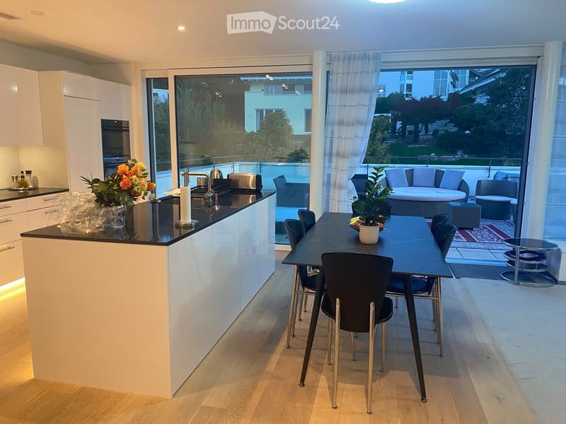 Modern kitchen with white cabinets, black countertops, and a central island. A dining table with chairs is adjacent, and a view through large windows shows a patio with a sofa and outdoor seating.