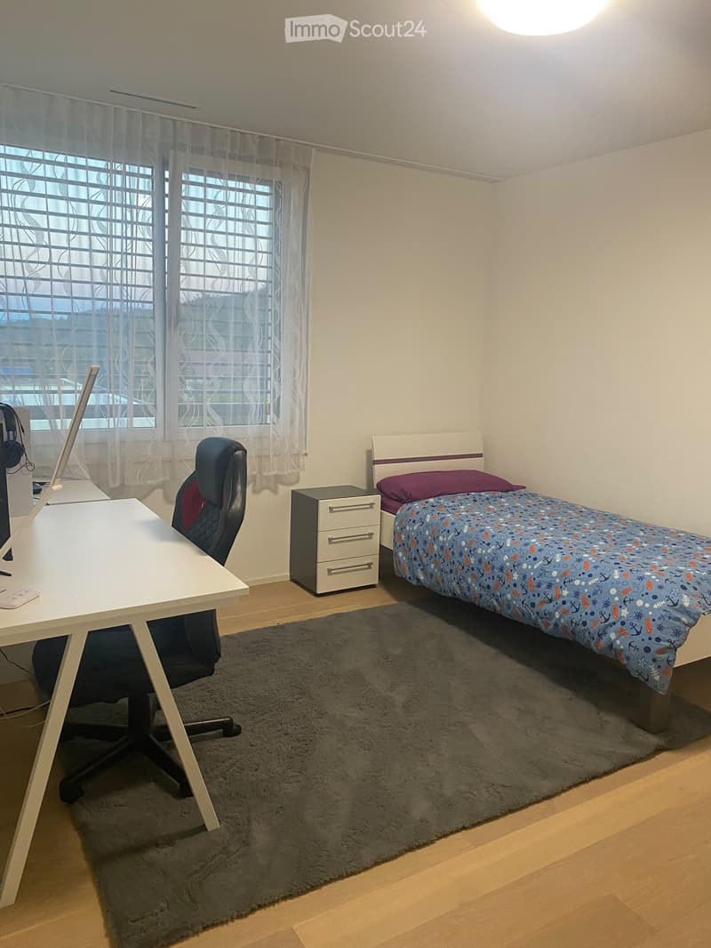 A small, well-lit bedroom with a single bed, desk, and chair. The bed has a colorful duvet and is positioned next to a window with sheer curtains. A desk with a laptop and a chair is placed near the window, and a small drawer unit is visible next to the b