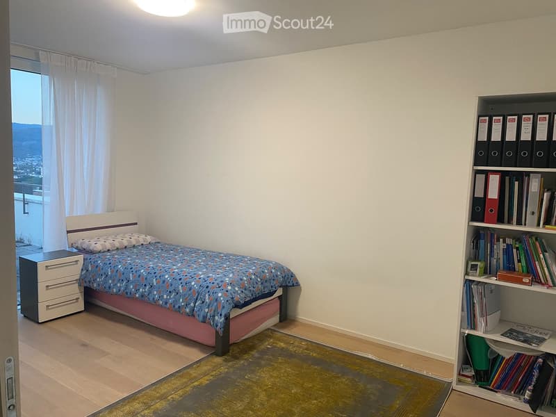 Cozy bedroom with a single bed, nightstand, bookshelf filled with books, and a scenic view through large windows.