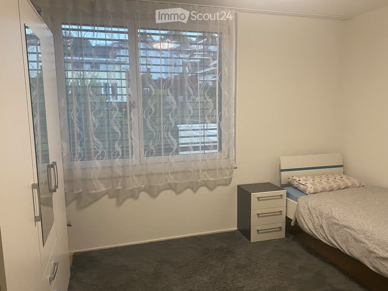 A tidy bedroom with a bed, nightstand, and a window with sheer curtains. The room features a grey carpet and white walls.