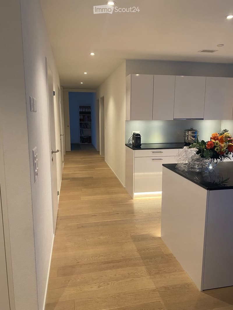A modern hallway with light wooden flooring, white walls, and recessed lighting. A small kitchen area is visible on the right with white cabinets, a black countertop, and a vase with flowers. A bookshelf is visible in the background.