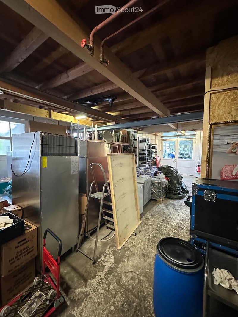 The image shows a cluttered attic or storage space with exposed wooden beams and pipes. Visible objects include a refrigerator, ladder, stool, speaker, barrel, handcart, and various boxes and cabinets.