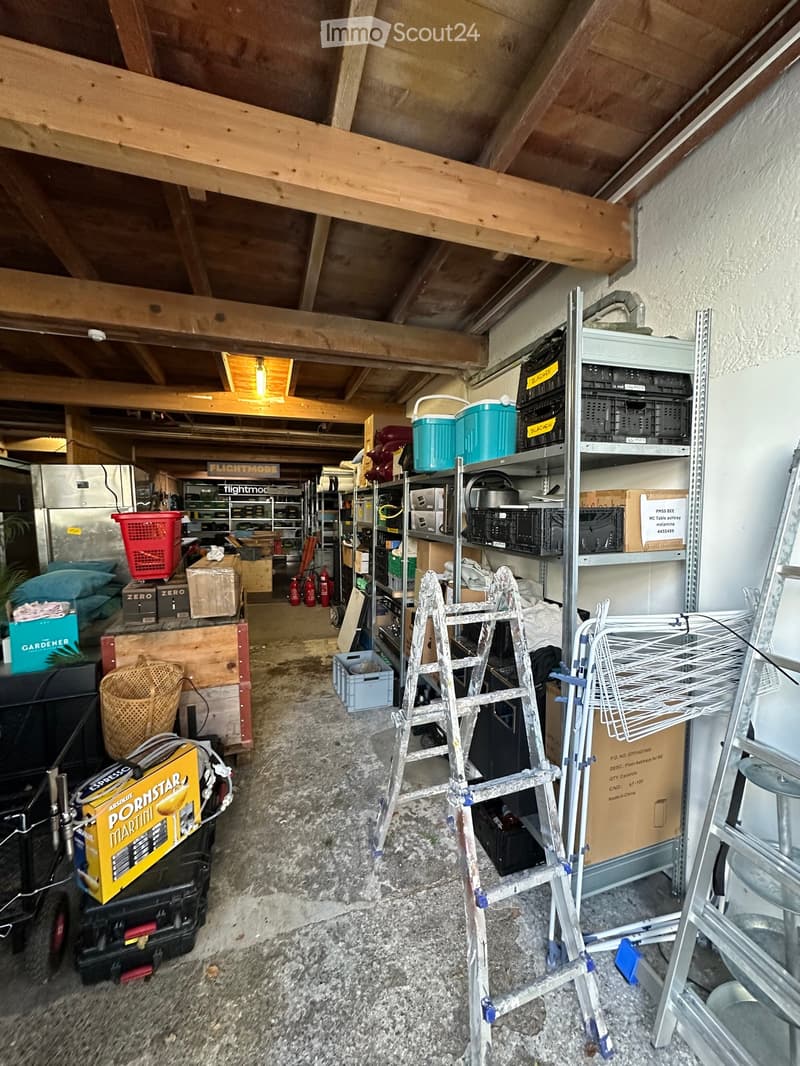The image shows an attic or storage area with exposed wooden beams and a slanted ceiling. The space contains various items including ladders, shelves filled with storage bins, a refrigerator, fire extinguishers, a wicker basket, and several boxes and cont