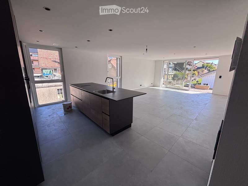 Modern kitchen with a central island, sleek black countertop, and a stainless steel sink. Large windows and sliding glass doors lead to an outdoor area.