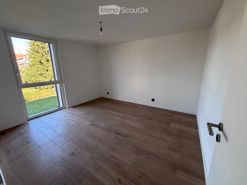 Empty room with wooden flooring, white walls, large window with view of greenery, and a door handle visible.