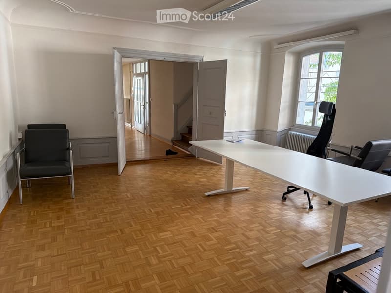 Office space with wooden herringbone flooring, white table, black office chairs, large window with view, open doorway leading to hallway.