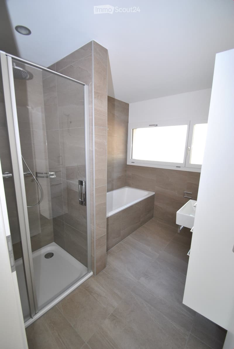 Modern bathroom with a bathtub, sink, and glass-enclosed shower stall. The walls are tiled in a warm, earthy tone, and the floor is covered with matching tiles. A window provides natural light.
