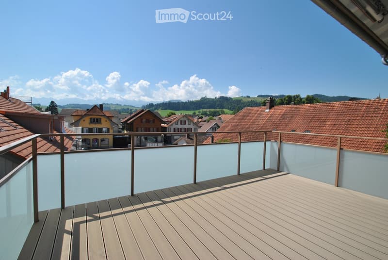 Balcony with wooden decking, glass railing, overlooking a village with red-tiled roofs and distant mountains.