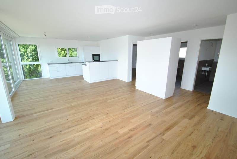 Bright, open-plan space with light wooden flooring, white walls, and large windows. The area includes a kitchen section with cabinets and a countertop, but no furniture or appliances are present.