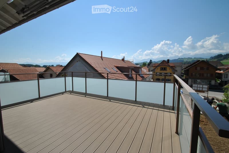 Wooden deck with glass railing, overlooking a village with red-tiled roofs and distant mountains.