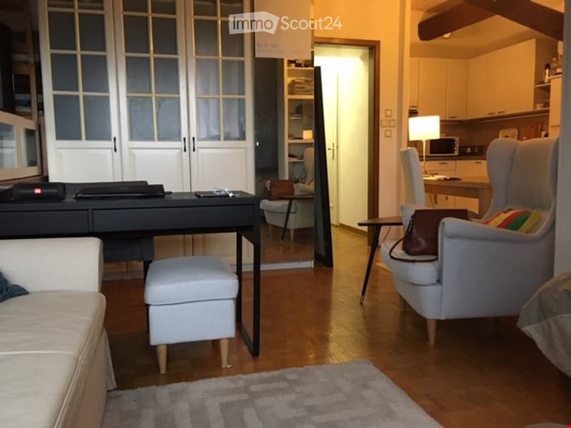 Bright, modern living space with hardwood flooring, a grey sofa, a small black desk with a laptop, a white armchair, a small round table, and a kitchen area with cabinets and a microwave.