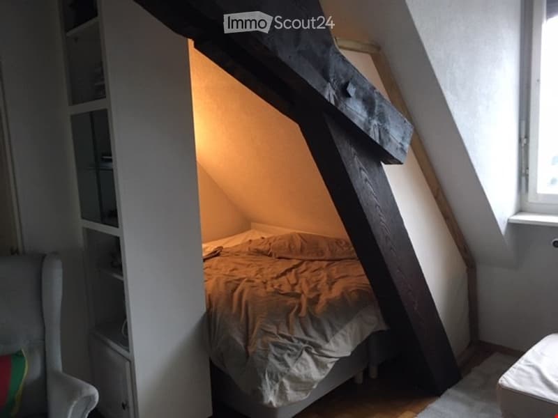 Cozy attic bedroom with slanted wooden beams, a single bed, a chair, and a bookshelf.