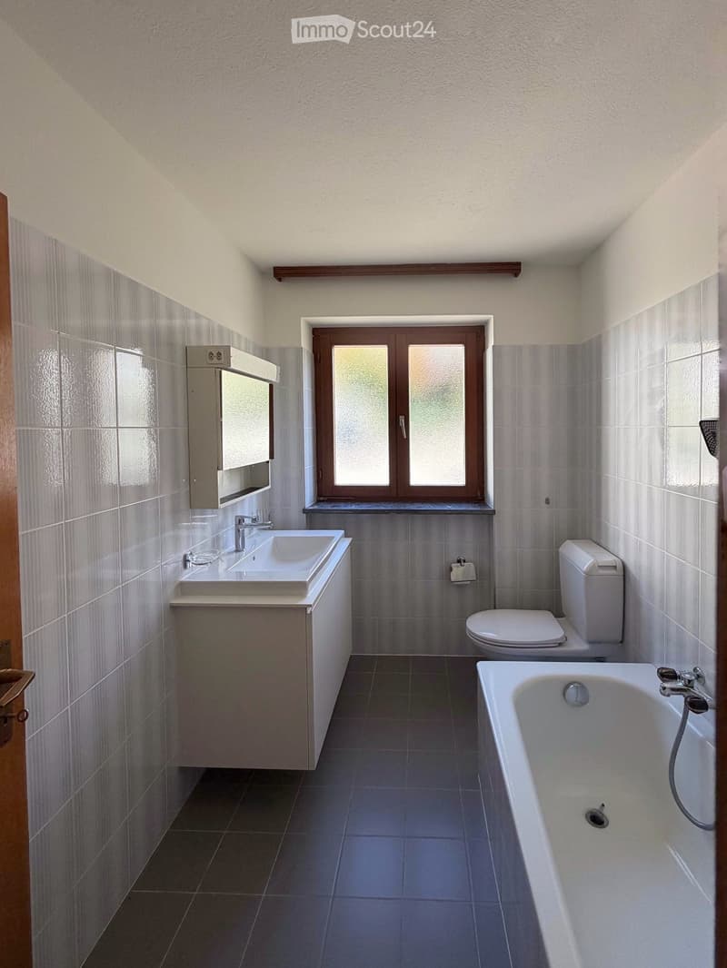 Modern bathroom with a white sink, toilet, bathtub, and mirror. Grey tiled walls and dark tiled floor, with a window providing natural light.