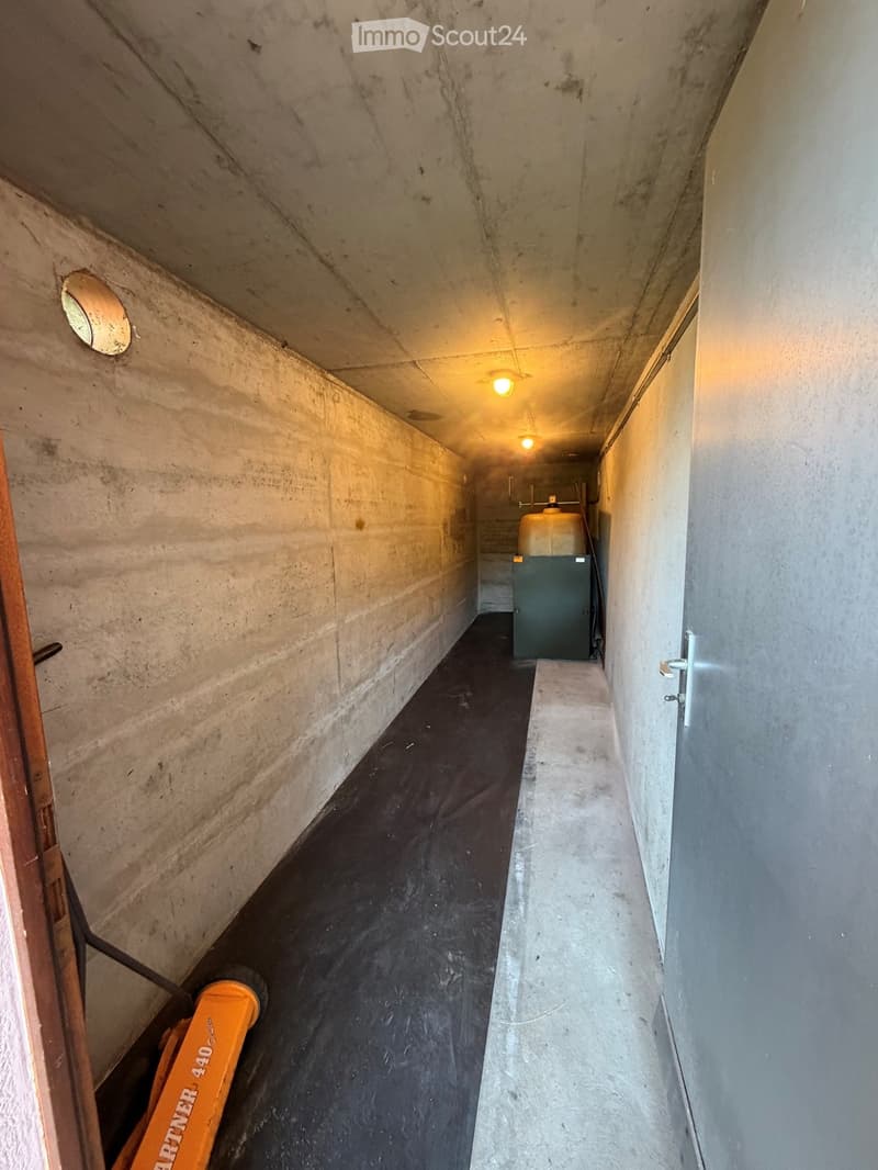 A narrow, dimly lit concrete corridor with a black floor, visible pipes, and a water tank in the background.