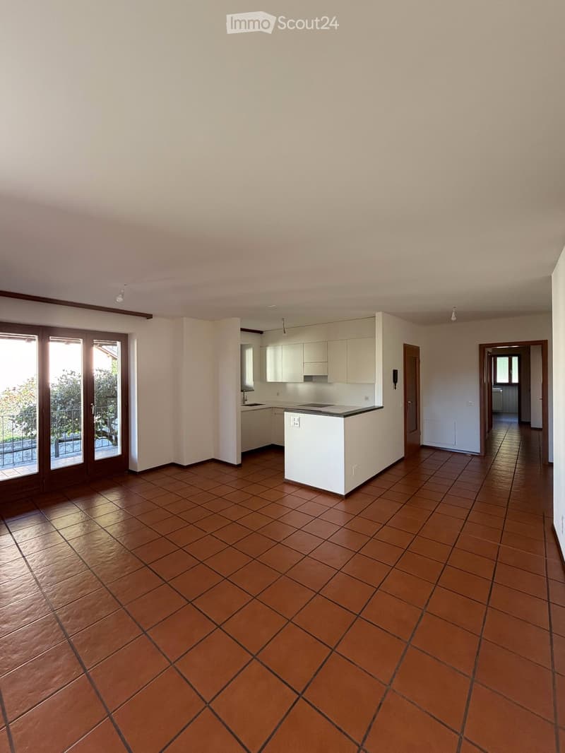 Empty room with tiled floor, white walls, and a small kitchen area visible in the background. The room has large sliding glass doors leading to a balcony.