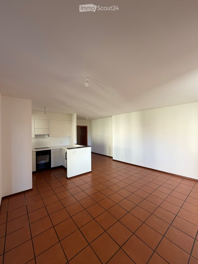 Empty room with tiled floor, white walls, and a small kitchen area with cabinets and appliances.