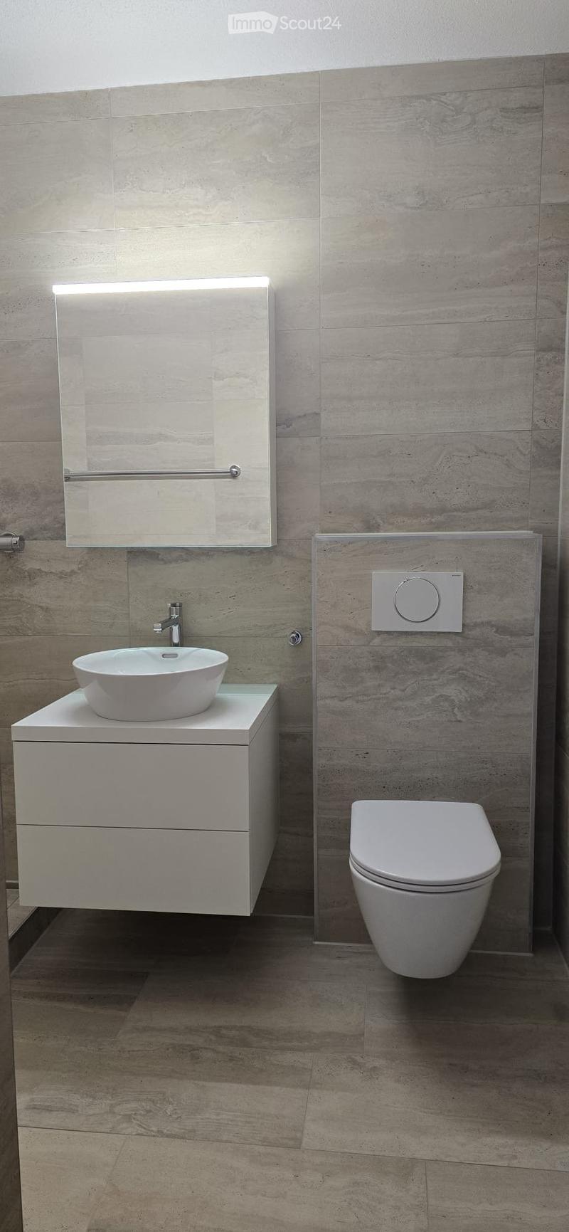 Modern bathroom with a white sink, toilet, and mirrored cabinet, featuring light grey tiled walls and a wooden floor.
