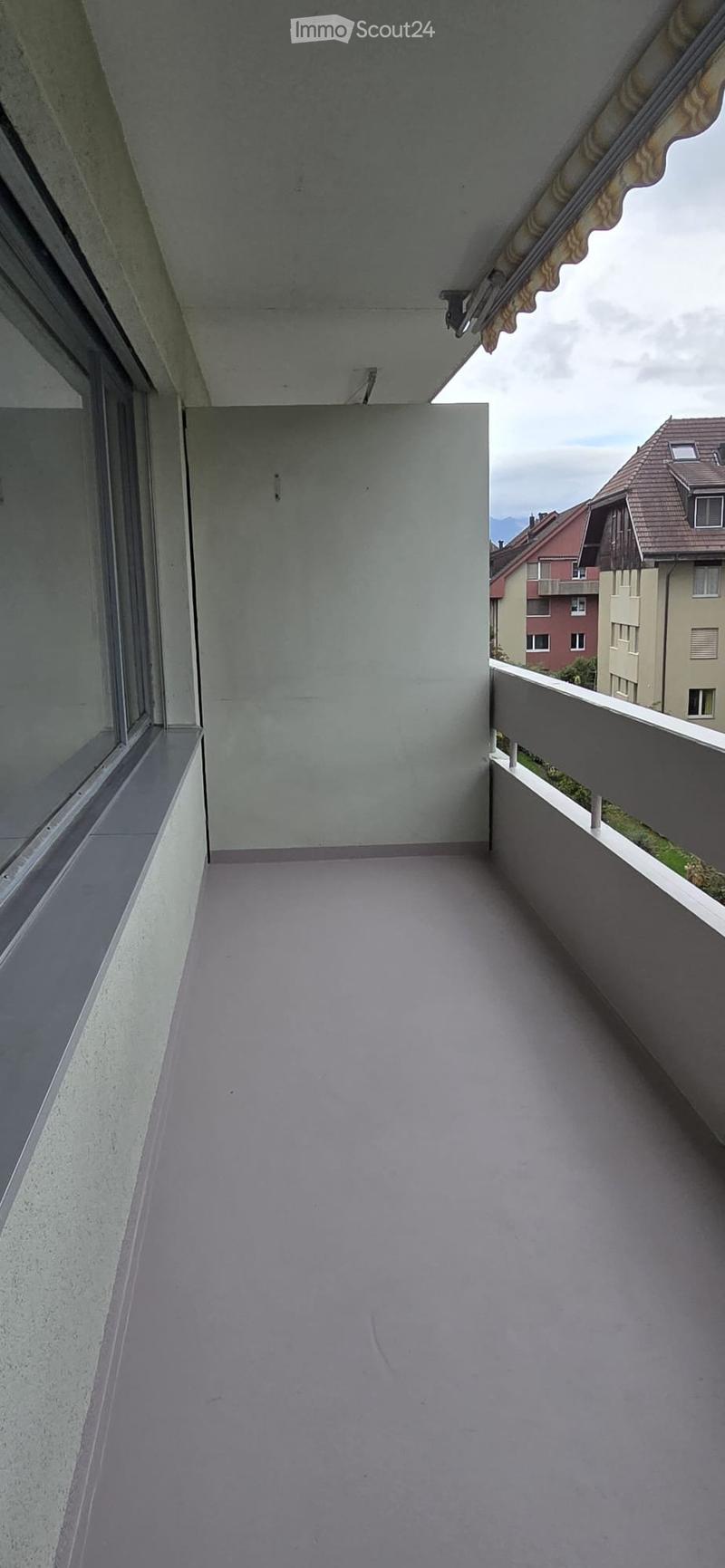 Empty balcony with grey concrete floor, white walls, and a metal railing, overlooking neighboring buildings and a cloudy sky.