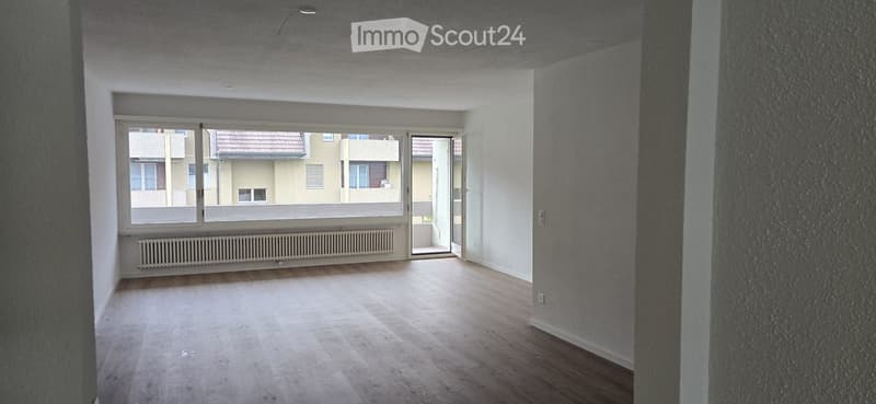 Empty room with wooden flooring, large window, radiator, and open doorway leading to a balcony.