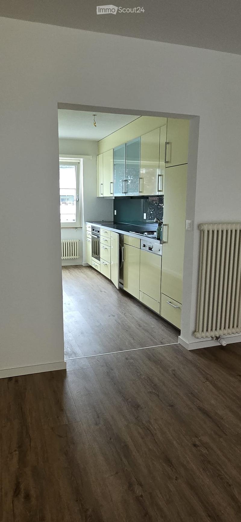 Empty room with wooden flooring, light-colored walls, and a small kitchen area visible in the background with cabinets and appliances.