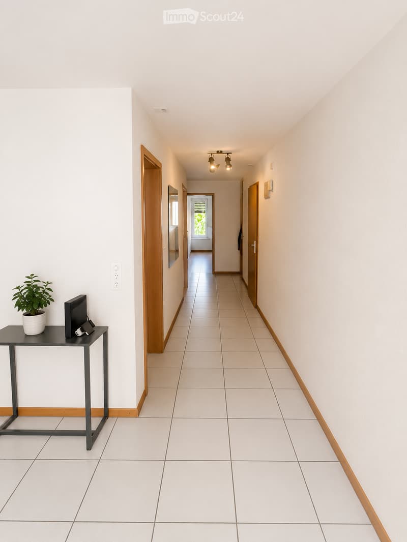 Clean, modern hallway with white walls, tiled floor, and wooden trim. Features recessed lighting, a small table with a plant and electronic devices on the left, and several doors leading to other rooms.