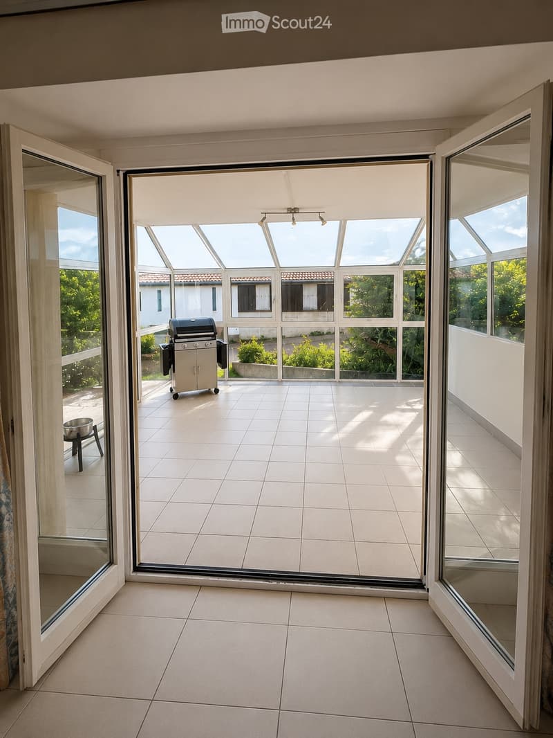 Bright, tiled balcony with large glass doors opening to a view of the garden and surrounding area. Features a grill and a small stool.