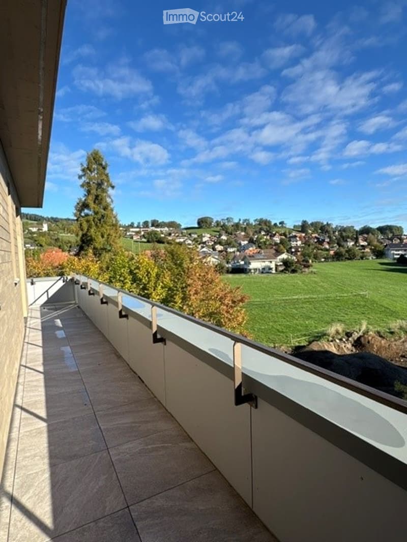 Balcony with a view of a green field, trees, and distant houses under a clear blue sky.