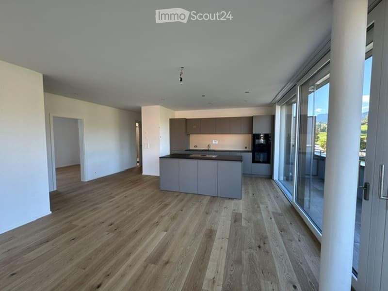 Modern, open-plan kitchen and living area with light grey cabinets, a central island, and large windows offering a scenic view.
