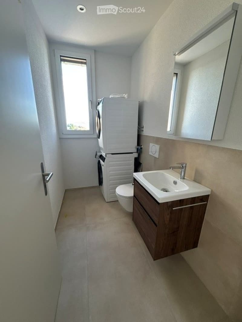 Modern bathroom with a white sink, mirror, washing machine, and toilet. Light-colored tiles and a window provide natural light.