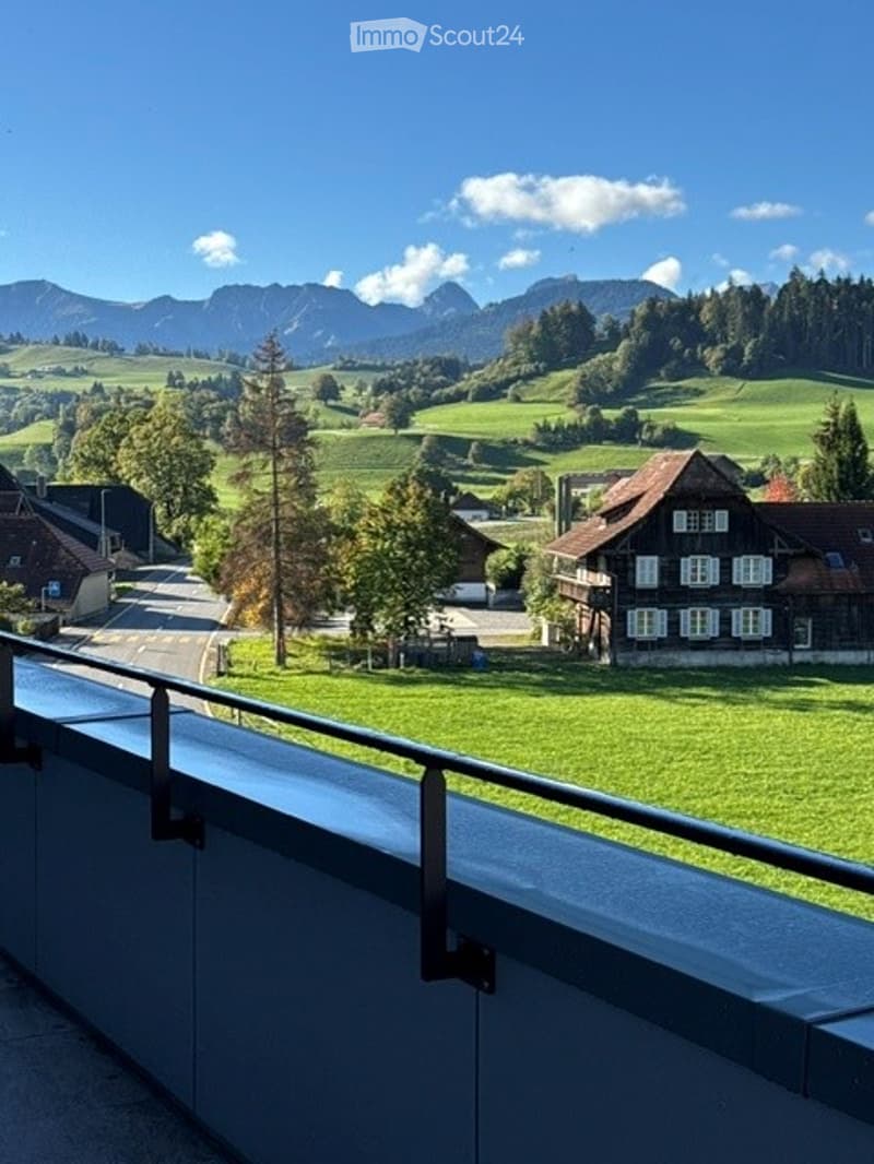 Balcony overlooking a scenic valley with green fields, trees, and distant mountains under a clear blue sky.
