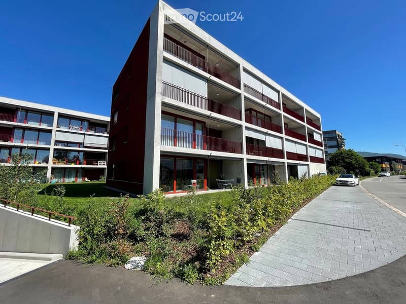 Modern multi-story residential building with red and white facade, balconies, and surrounding greenery.