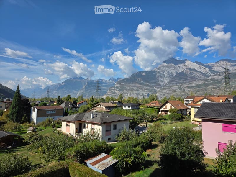 A residential area with multiple houses surrounded by greenery, with a mountain range in the background under a clear blue sky.