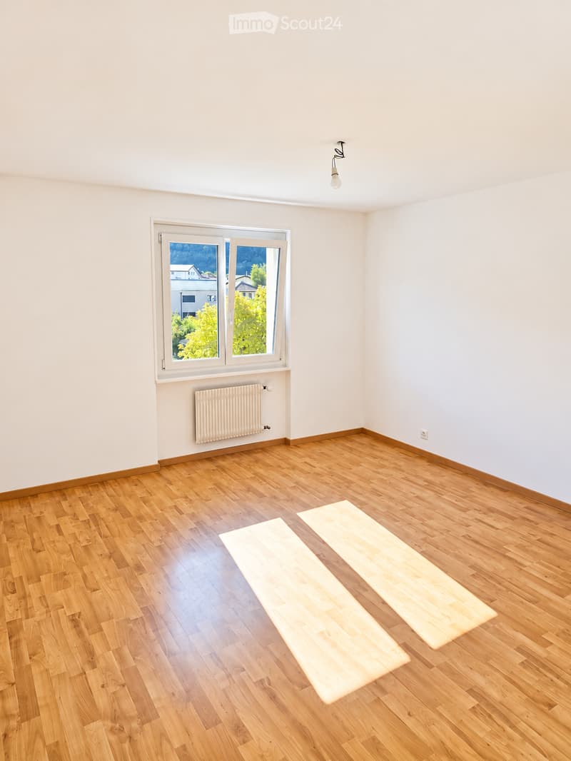 Empty room with wooden flooring, white walls, a window with a view of greenery, a radiator, and a ceiling light fixture.