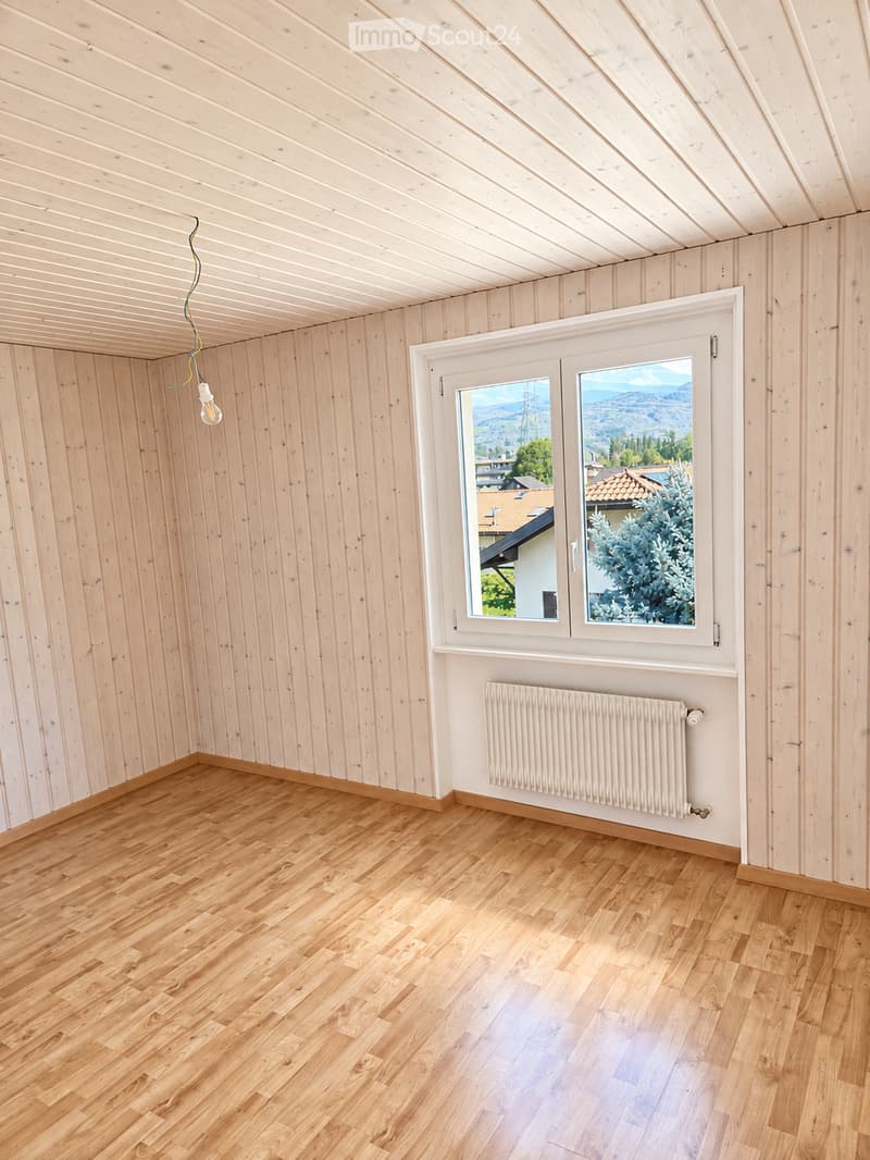 Empty room with wooden walls, wooden floor, large window with scenic view, radiator, and exposed wooden ceiling beams.