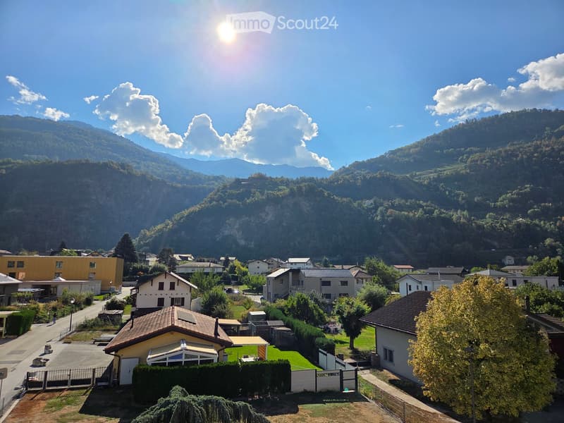 A scenic view of a residential area nestled in a valley with mountains in the background, featuring houses, trees, and a clear blue sky.
