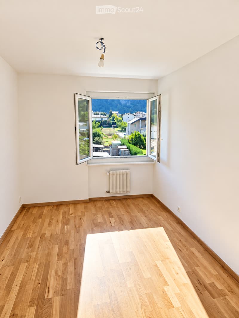 Empty room with wooden flooring, white walls, large window with scenic view, radiator, and a single light fixture.