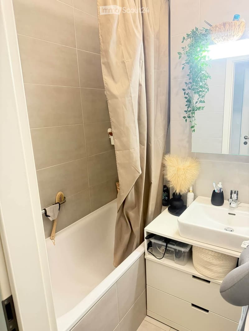 Modern bathroom with a bathtub, sink, mirror, and various toiletries. Beige tiles and a window with a decorative plant visible.