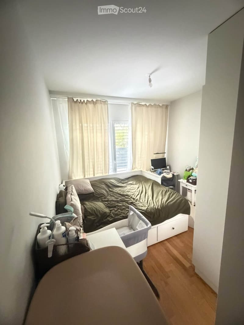 A small bedroom with a bed, baby cot, desk, and chair. The room features wooden flooring, white walls, and large windows with curtains.