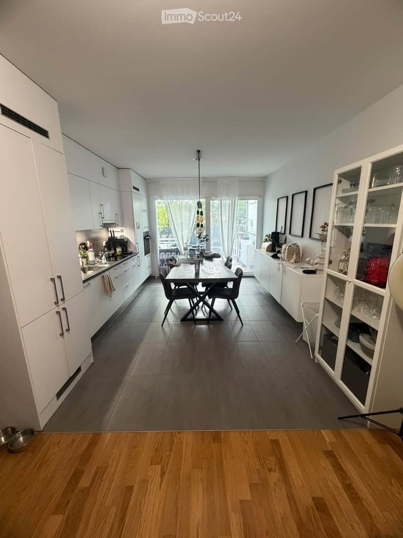Open-plan kitchen and dining area with white cabinets, a dark dining table with chairs, and a window with curtains leading to a balcony.