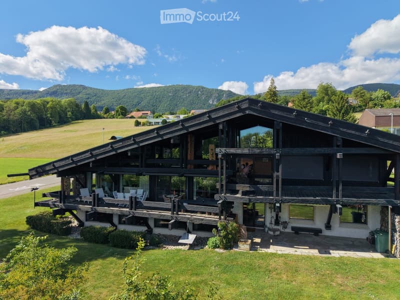 Two-story wooden house with large windows and a spacious wooden deck, surrounded by a well-maintained lawn and scenic mountain backdrop.