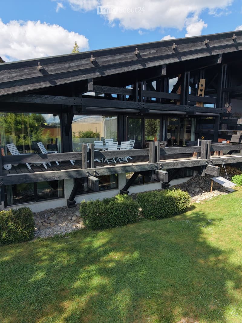 Modern two-story wooden house with large windows, a spacious wooden deck, and outdoor seating area.
