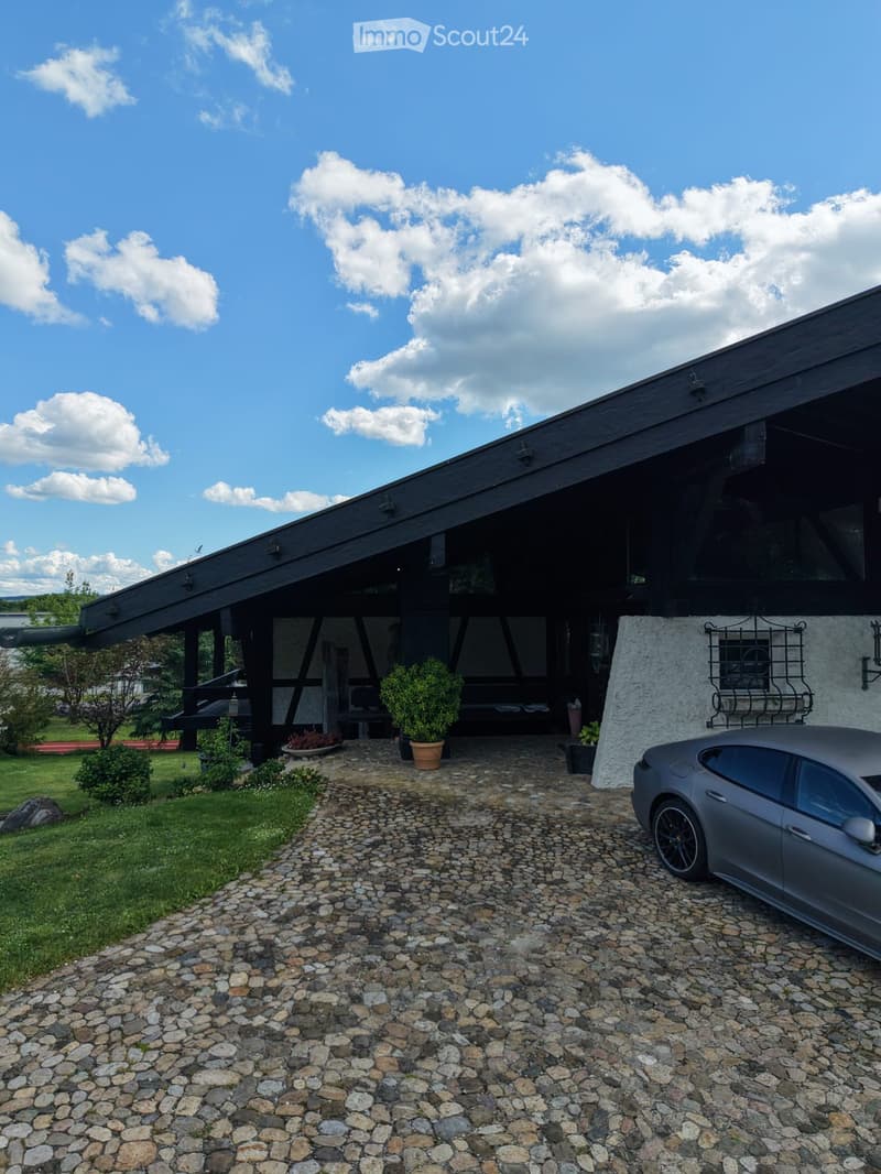 Modern two-story house with a dark wooden roof and large windows, featuring a stone wall, potted plants, and a parked car on a cobblestone driveway.