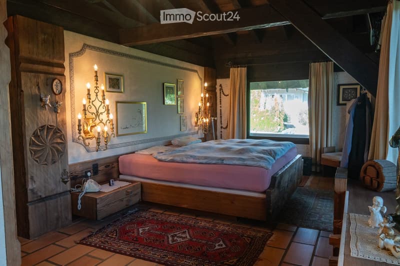 Cozy bedroom with wooden beams, a large bed with pink bedding, nightstand with a telephone, wall-mounted chandelier, framed artwork, and a large window with curtains.