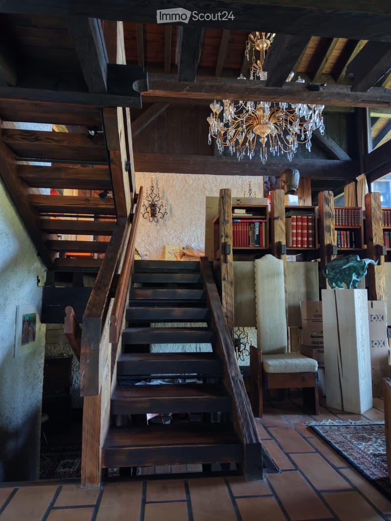 A spacious, well-lit room with wooden beams and a high ceiling. Features a wooden staircase leading to an upper level, a chandelier, bookshelves filled with books, a chair, and a small table.