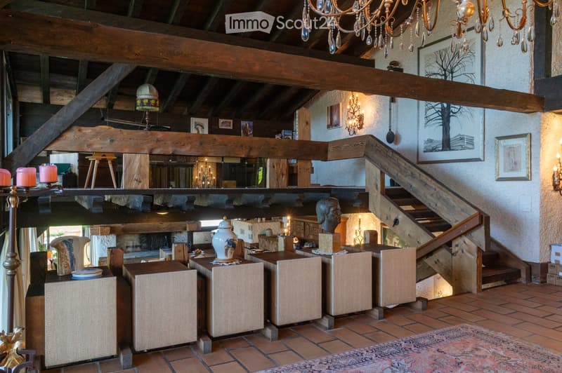 A spacious, well-lit living area with exposed wooden beams, a large staircase, and a cozy seating arrangement. The room features a mix of modern and rustic decor, including a large chandelier, wall-mounted lamps, and various decorative items on tables and