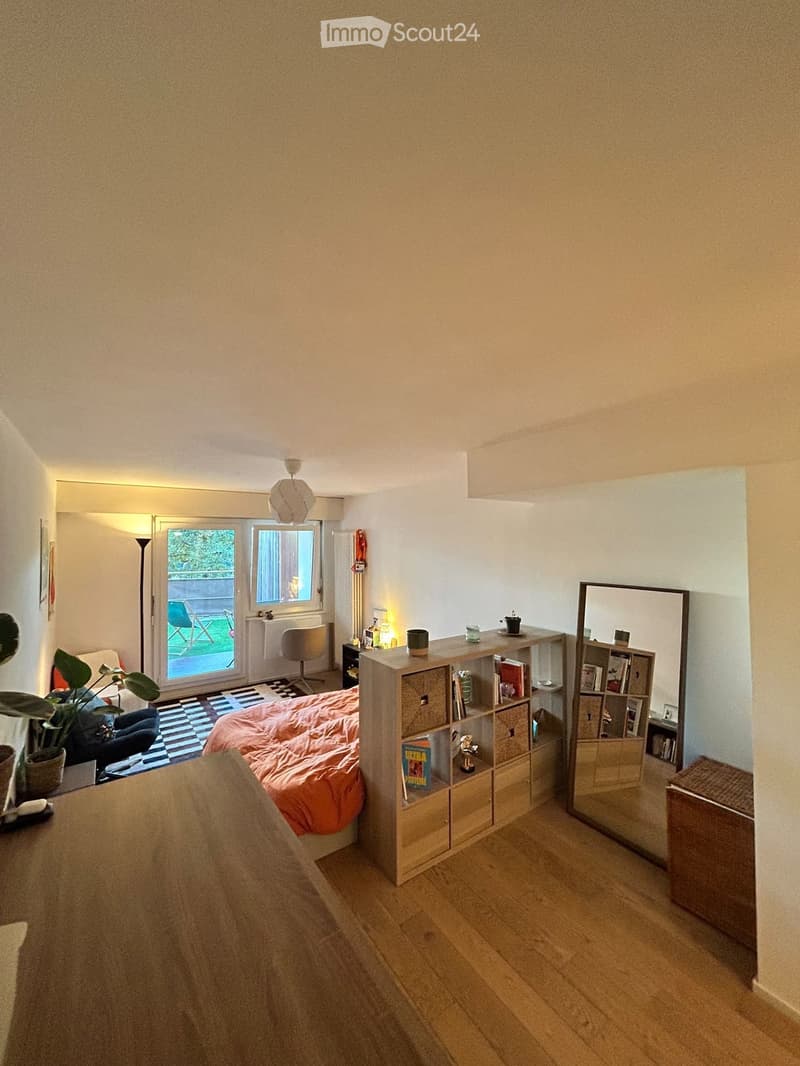 Cozy bedroom with a bed covered in an orange blanket, a small sofa, a wooden bookshelf, a mirror, and a desk area with a chair and lamp.