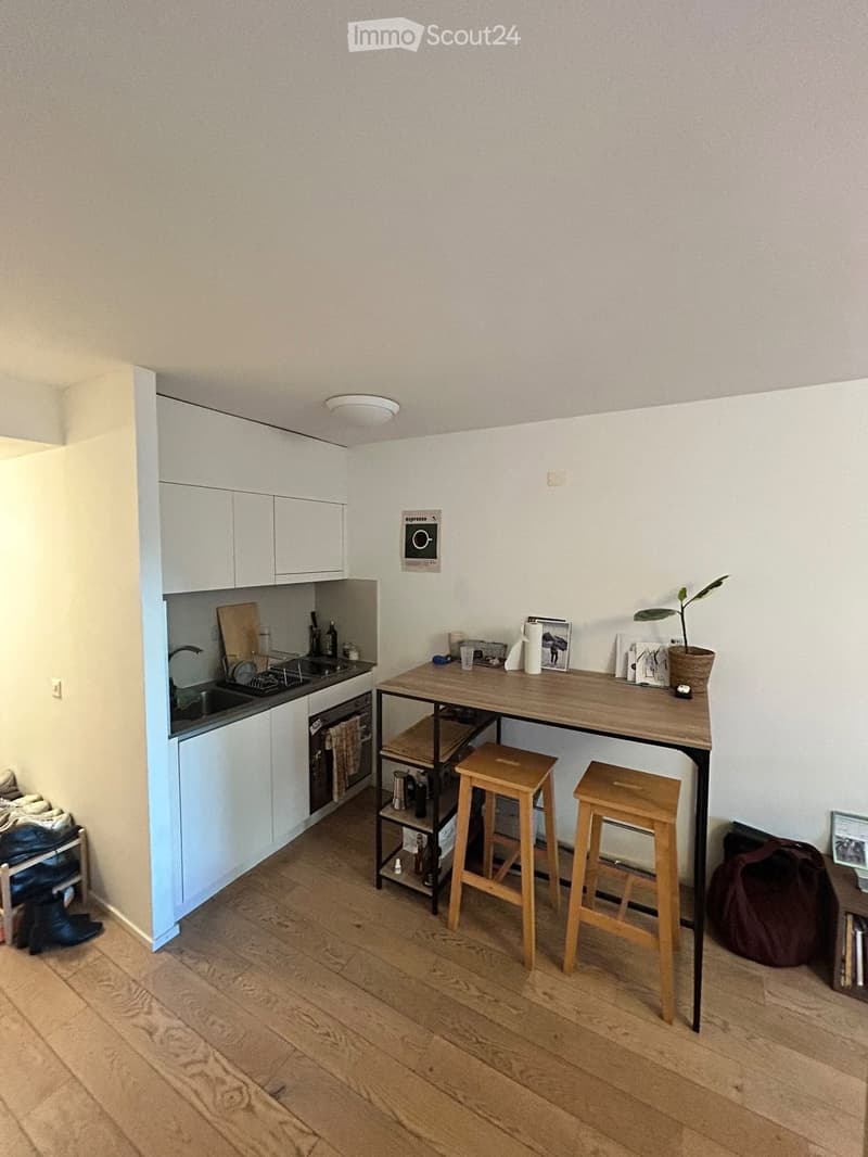 Modern kitchen with white cabinets, wooden floor, sink, oven, and a small dining area with a table and two stools.
