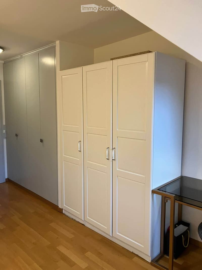 Empty room with light-colored wooden flooring and white built-in wardrobes with multiple doors and handles. A small glass-top table with a metallic frame is visible in the corner.